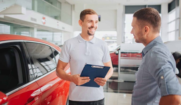 Young bearded man stands in front of customer and smiles. He holds plastic tablet with both hands. People are in front of red and beautfiul car. Customer is serious